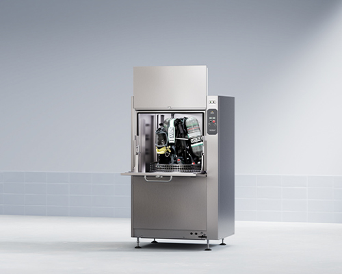 Stainless steel industrial dishwasher in clean room, door open showing racks and SCBA gear inside.